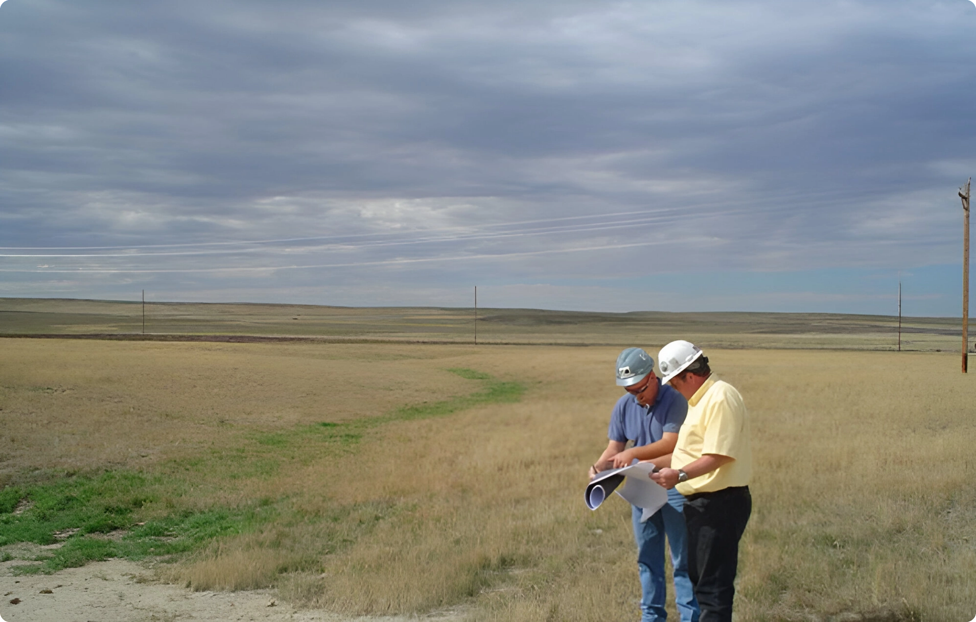 Two men in hard hats examine architectural plans in an open grassy field under a cloudy sky.