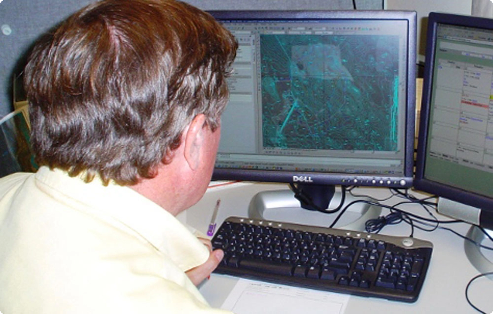 Person with short hair sits at a desk, viewing aerial maps on dual computer monitors. A keyboard and paper are on the desk.