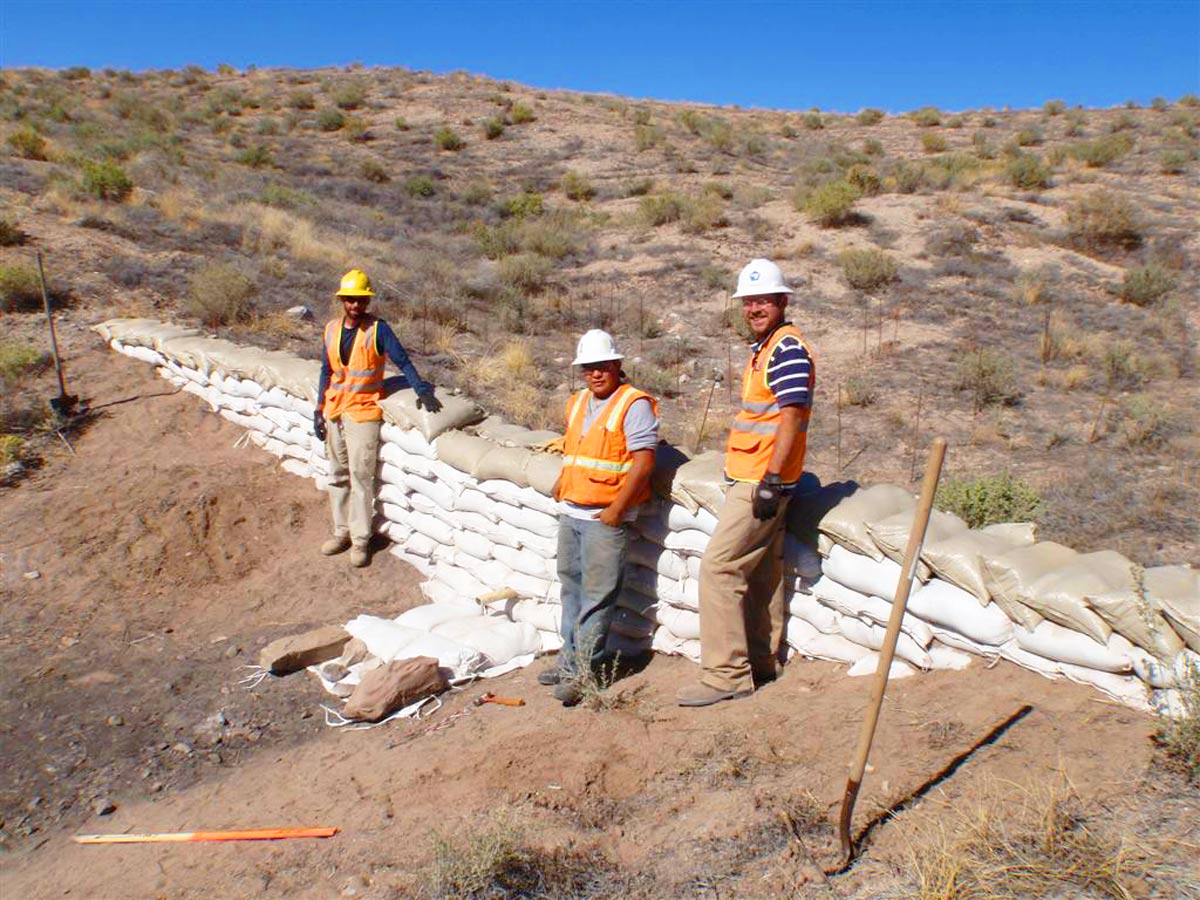 Three construction workers in vests and helmets stand beside a wall of sandbags on a dry, hilly terrain under a clear blue sky. A shovel is on the ground nearby.