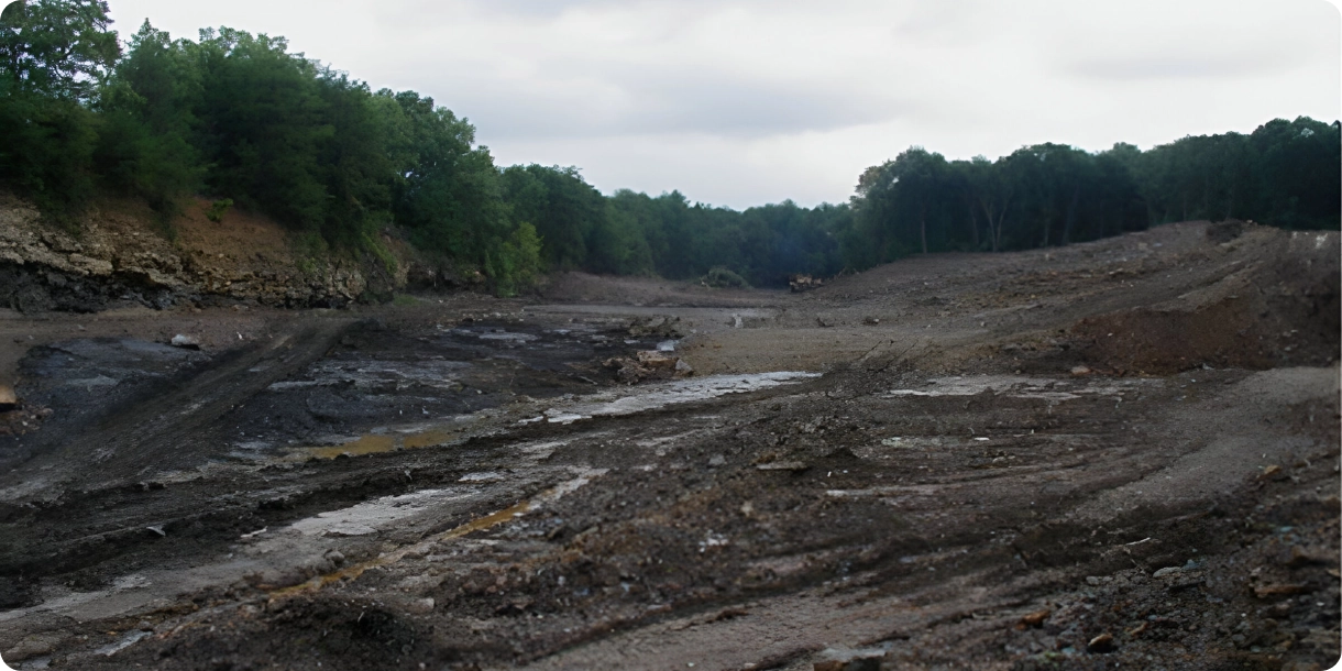 Cleared land area with rugged terrain and exposed soil, surrounded by trees under an overcast sky.