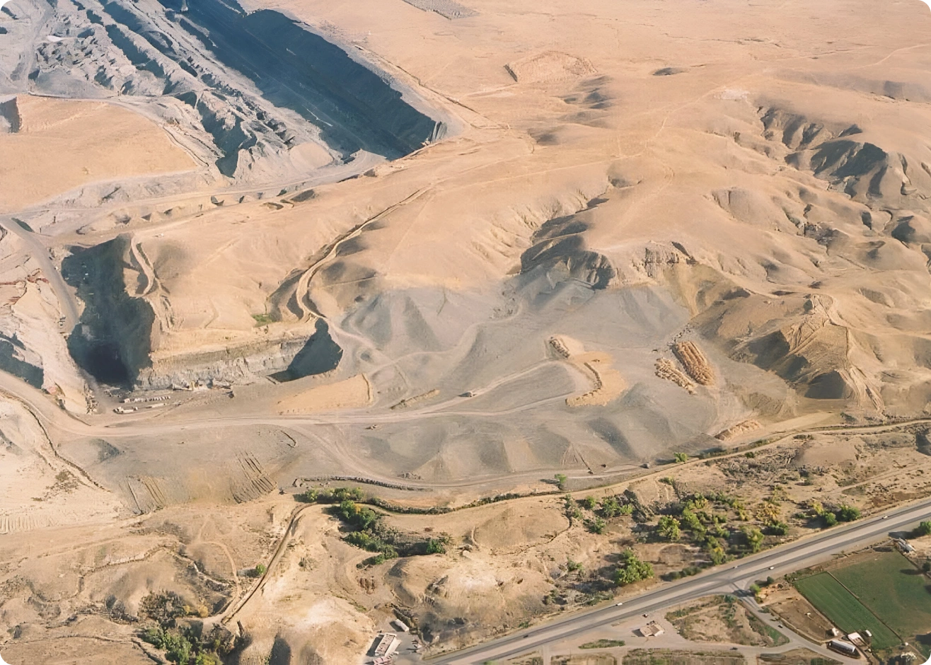 Aerial view of a desert landscape with hills, a quarry, and winding roads. Green patches and roads are visible at the bottom of the image.