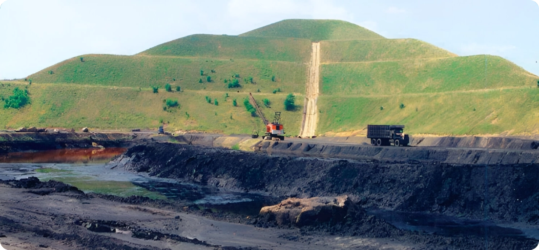 A mining site with heavy machinery, including an excavator and a truck, working near a large, grassy hill under a clear sky.