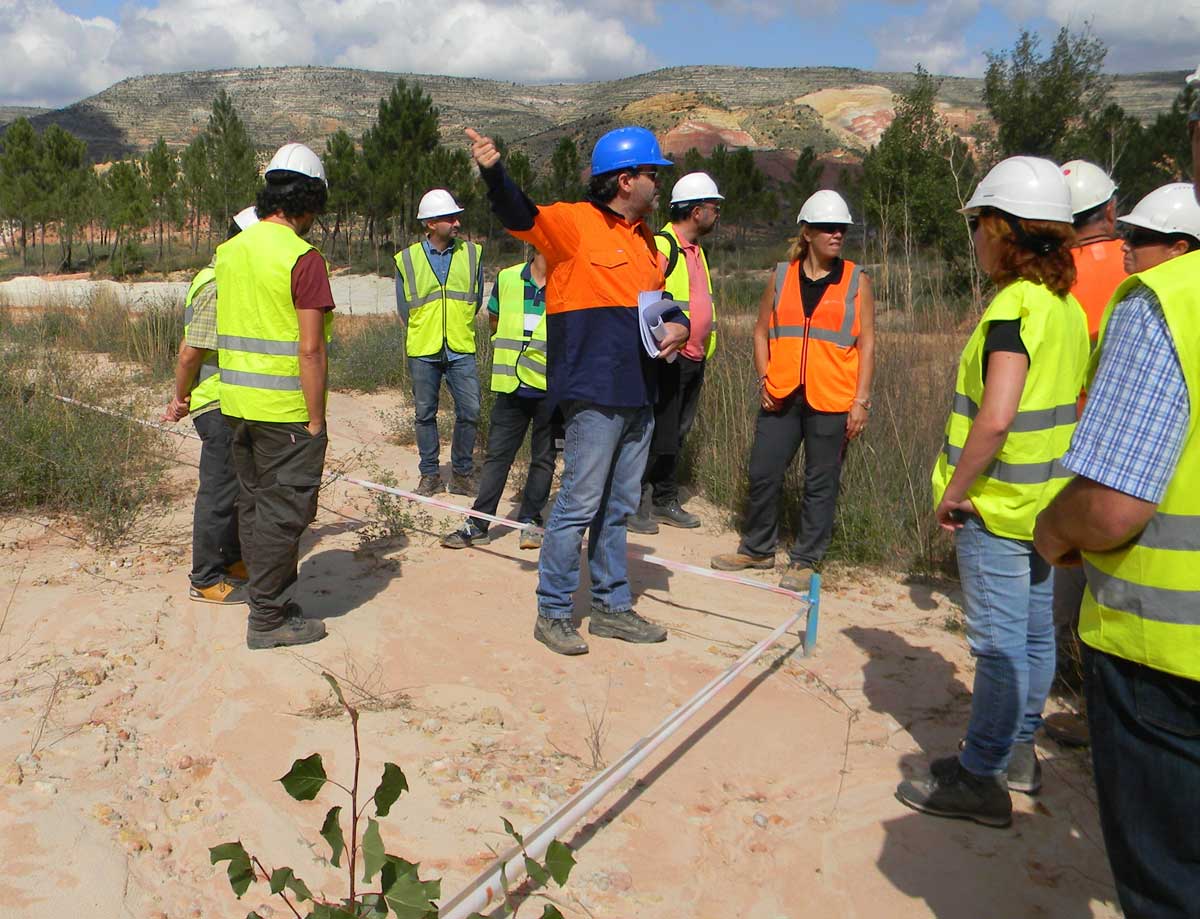 A group of people wearing hard hats and safety vests stand outdoors on sandy ground, with one person gesturing and speaking. Trees and hills are visible in the background.