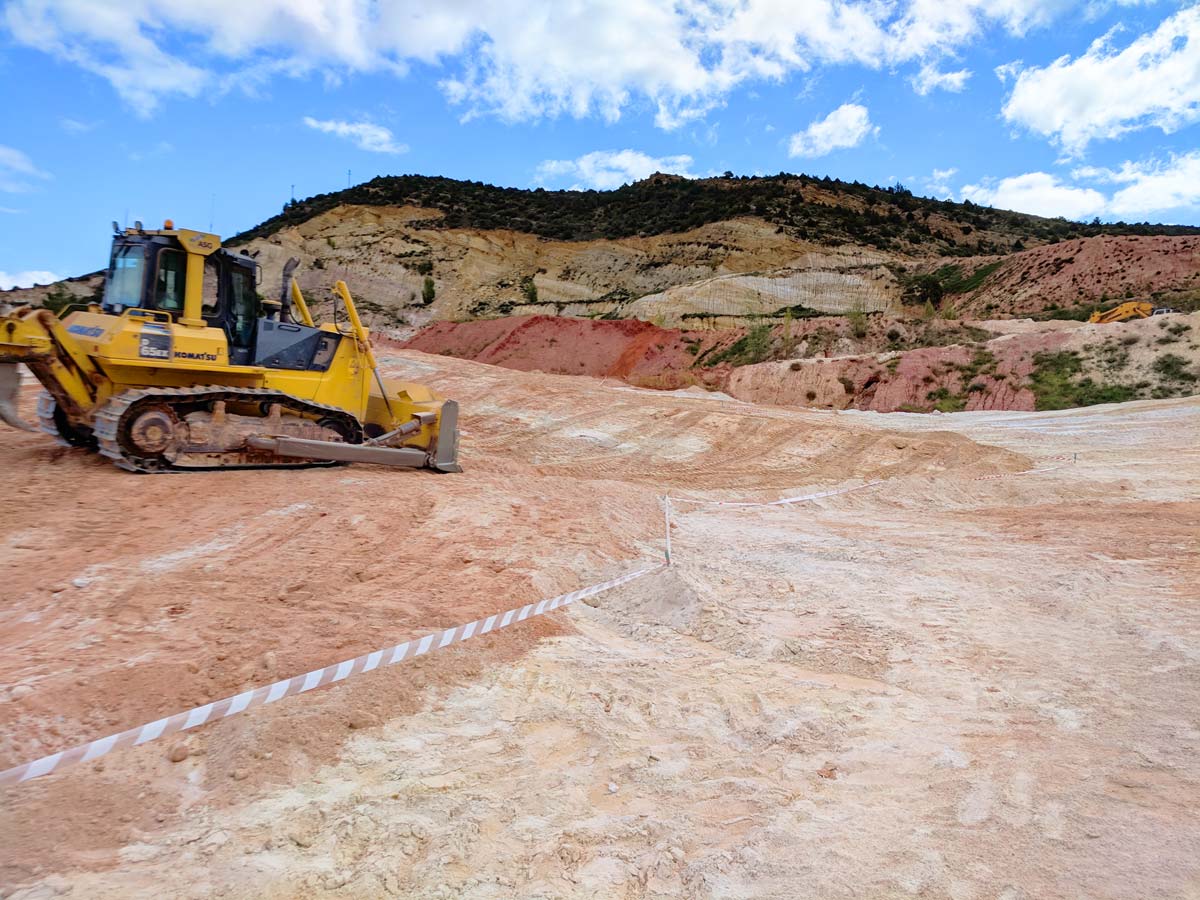 A yellow bulldozer on a sandy construction site with marked boundaries, against a backdrop of hills and a partly cloudy sky.