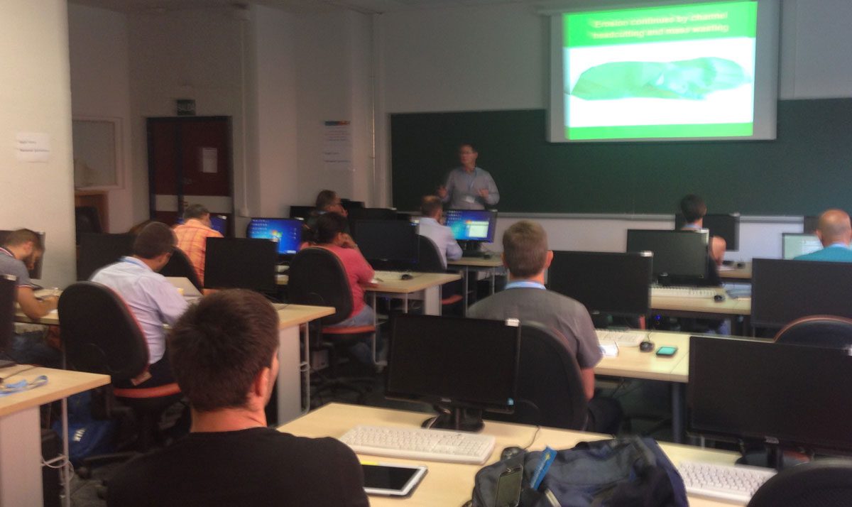 A classroom with students seated at desks with computers. An instructor stands at the front near a projection screen displaying a green presentation slide.