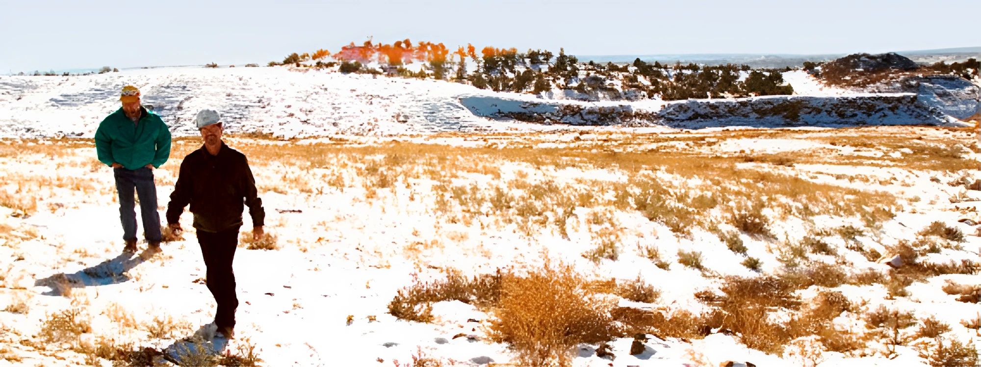 Two people walking through a snow-dusted, grassy landscape with distant trees and hills under a clear sky.