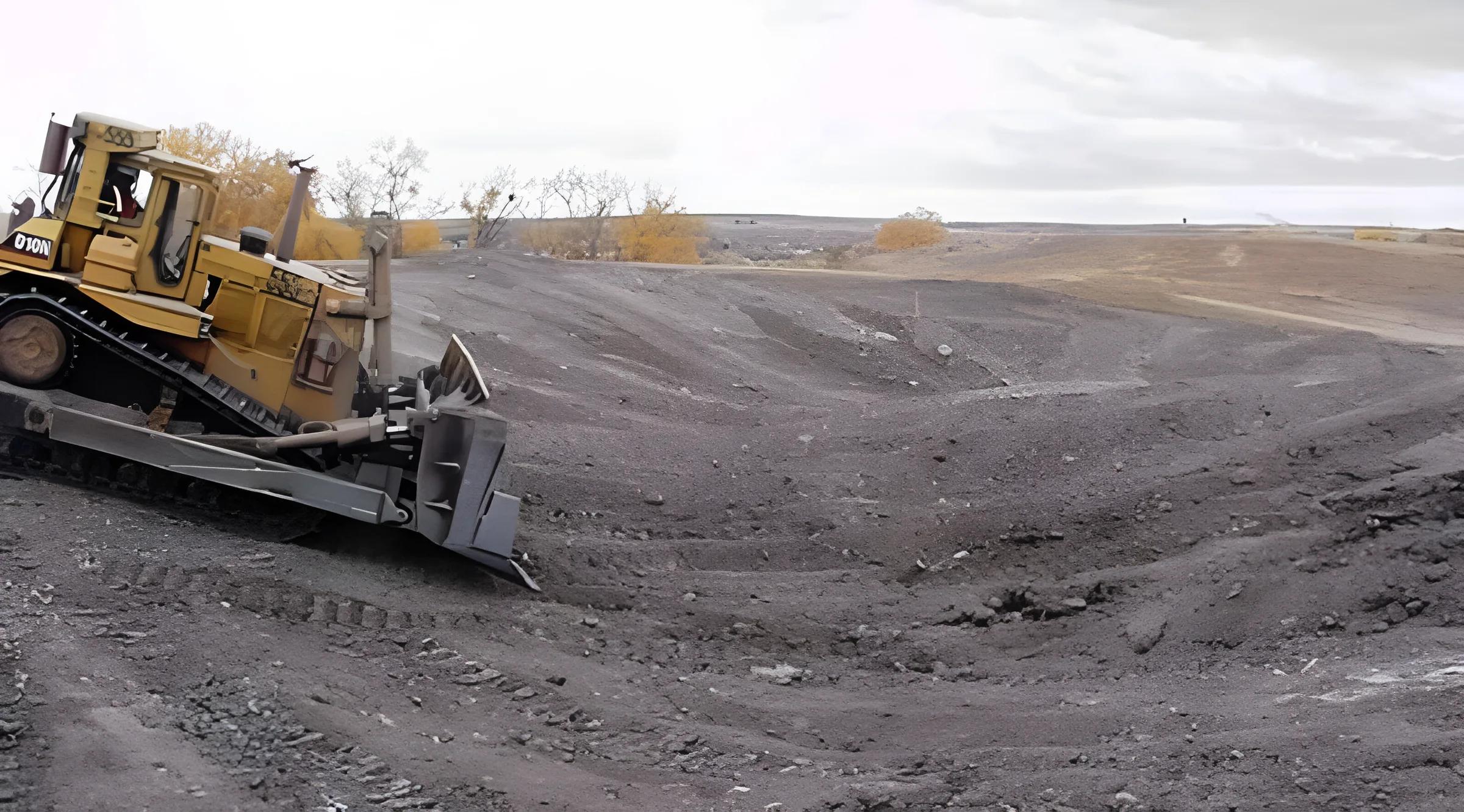 A bulldozer moves across a barren, grey landscape, pushing earth. The sky is overcast and sparse trees are visible in the background.