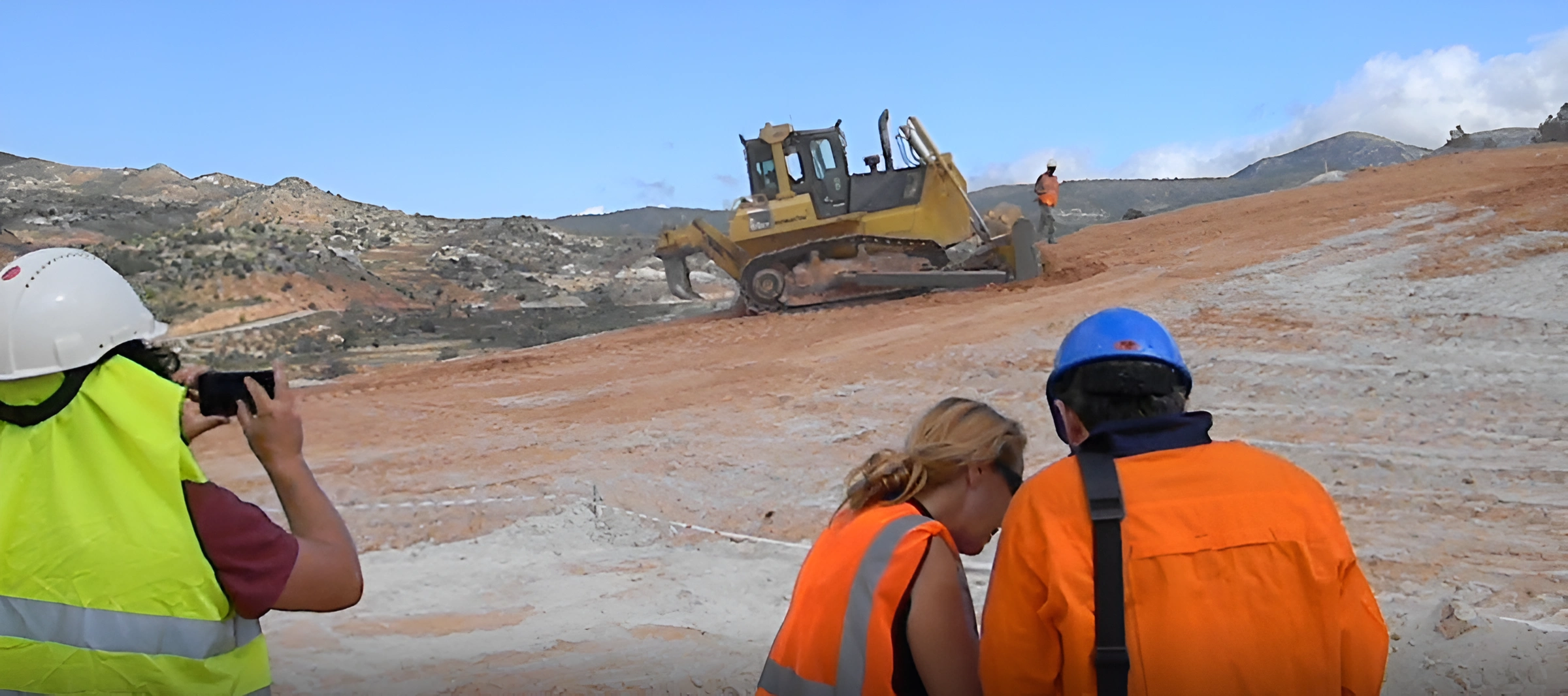 Construction workers wearing safety gear observe a bulldozer moving earth on a hilly site, with a mountainous landscape in the background.