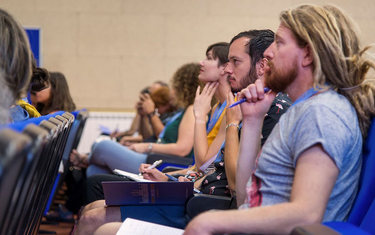 A group of people seated in a row, attentively listening during an indoor conference or seminar.