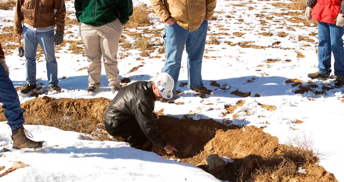 A group of people in jackets and gloves gather around a small hole in snowy terrain, observing a person in a helmet kneeling and examining the ground.